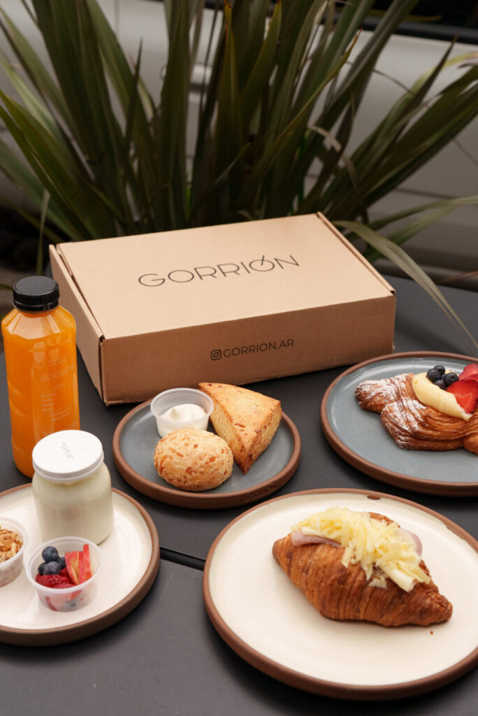 Brunch servido en platos de cerámica junto a la caja Gorrión: croissant, danesa, chipá, scon, yogur y jugo
