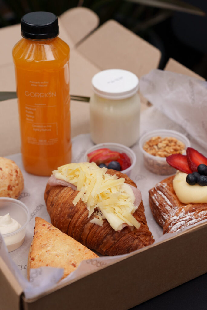 Caja de desayuno Gorrión con croissant de jamón y queso, danesa con frutos rojos, scon, chipá, yogur con granola y jugo prensado en frío