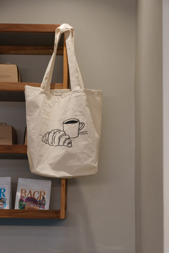 Tote bag de tela Gorrión con ilustración de café y croissant, colgada en el estante del local